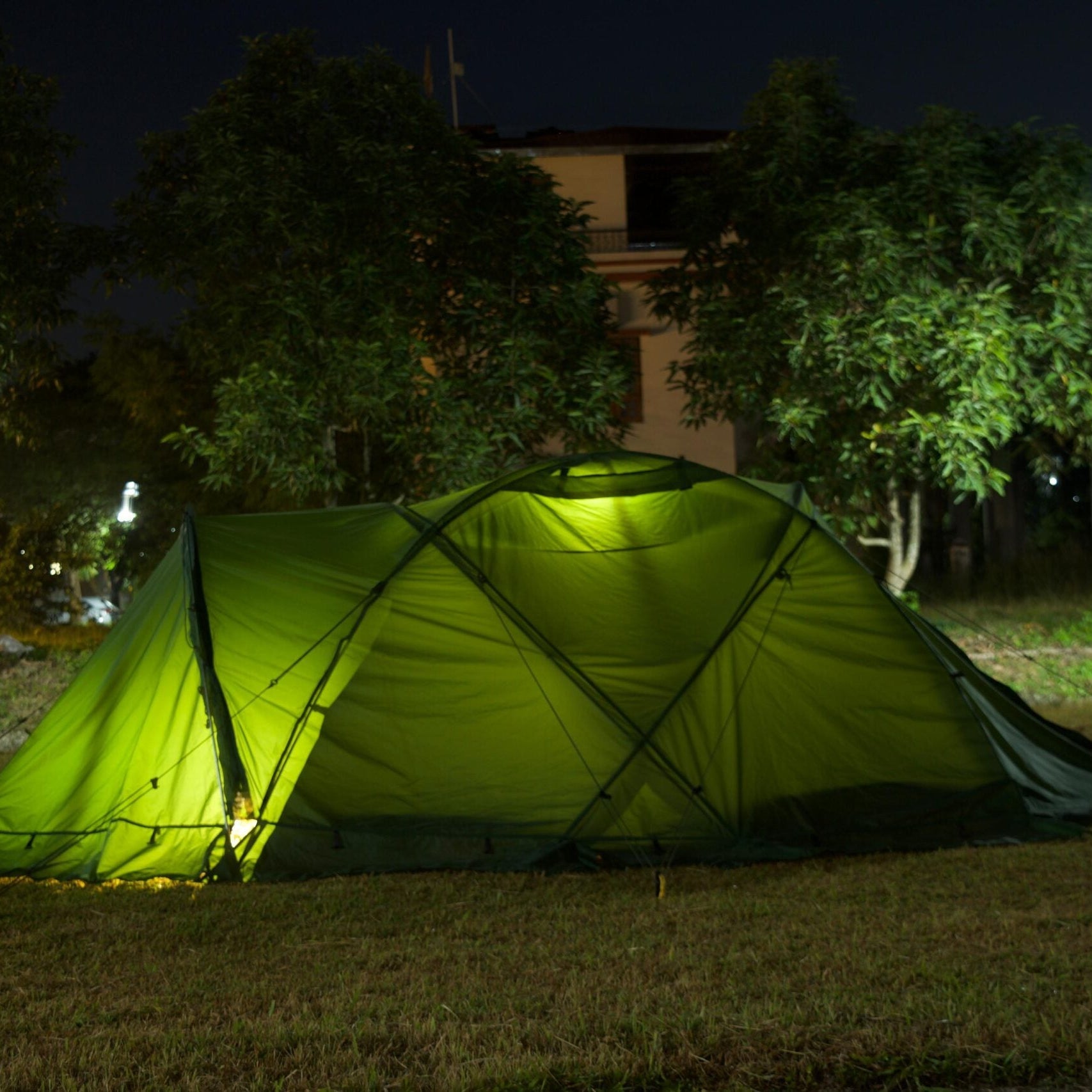 Green tent illuminated at night in a grassy area with trees and buildings in the background